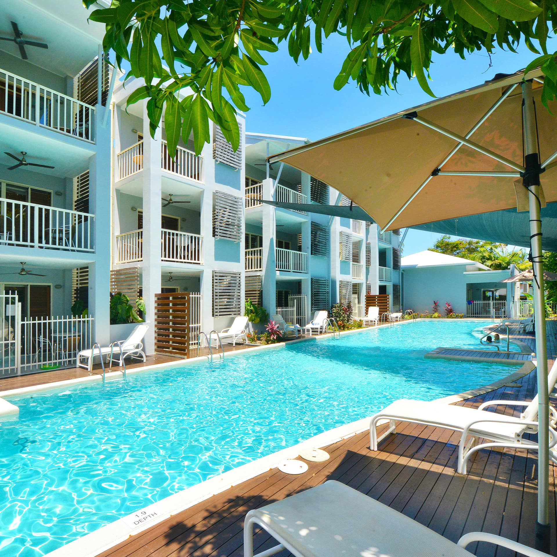 A long pool extends along the side of an apartment complex with balconies on each level, lounge chairs on wood patio space.