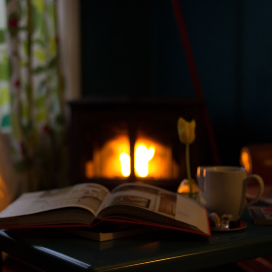 Cosy den with open book, coffee cup and flower on a table in the foreground of a blurred burning fireplace