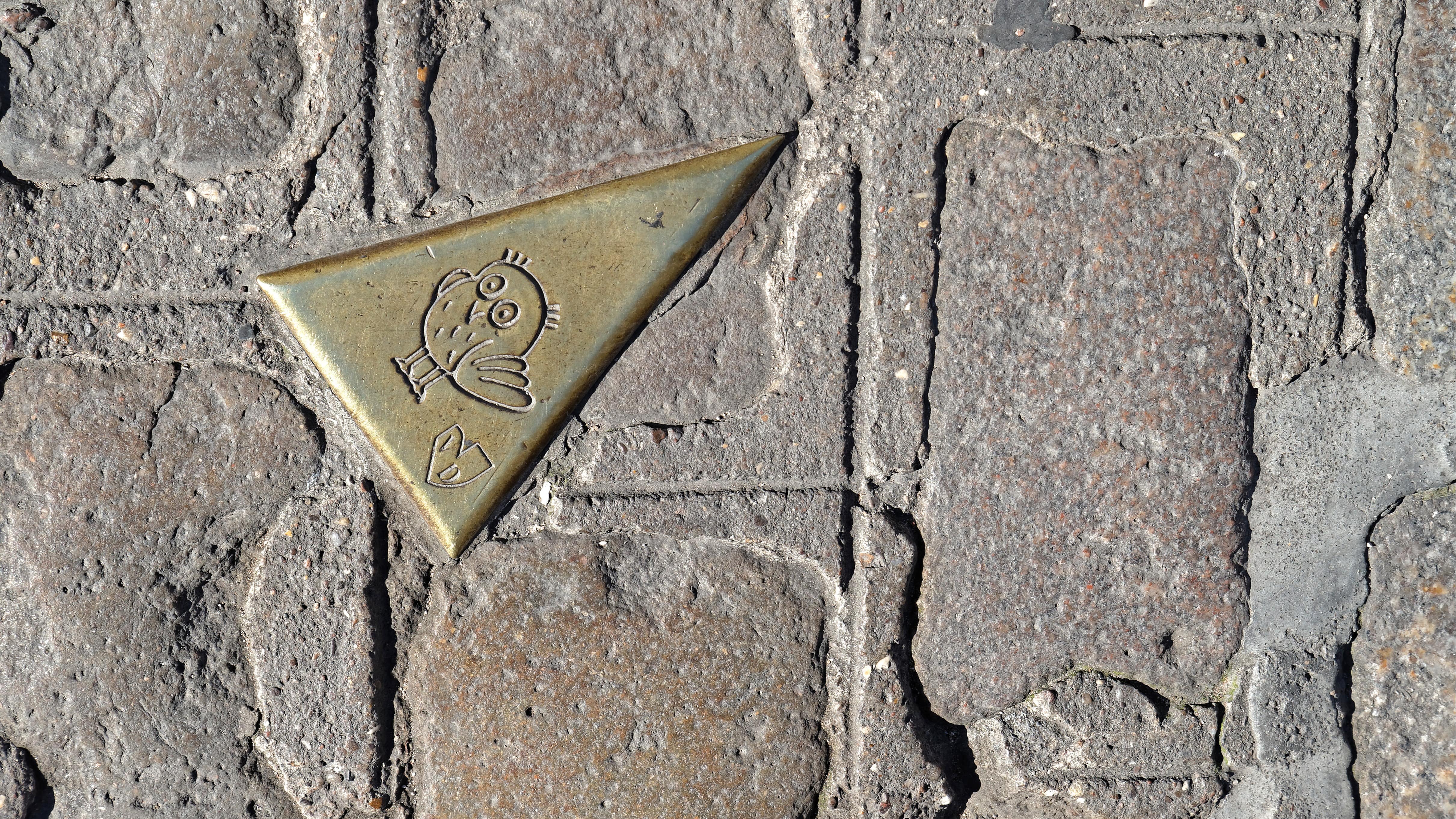 Par terre, entre les pavés, vue sur une plaque dorée triangulaire représentant une chouette, faisant partie du parcours du même nom
