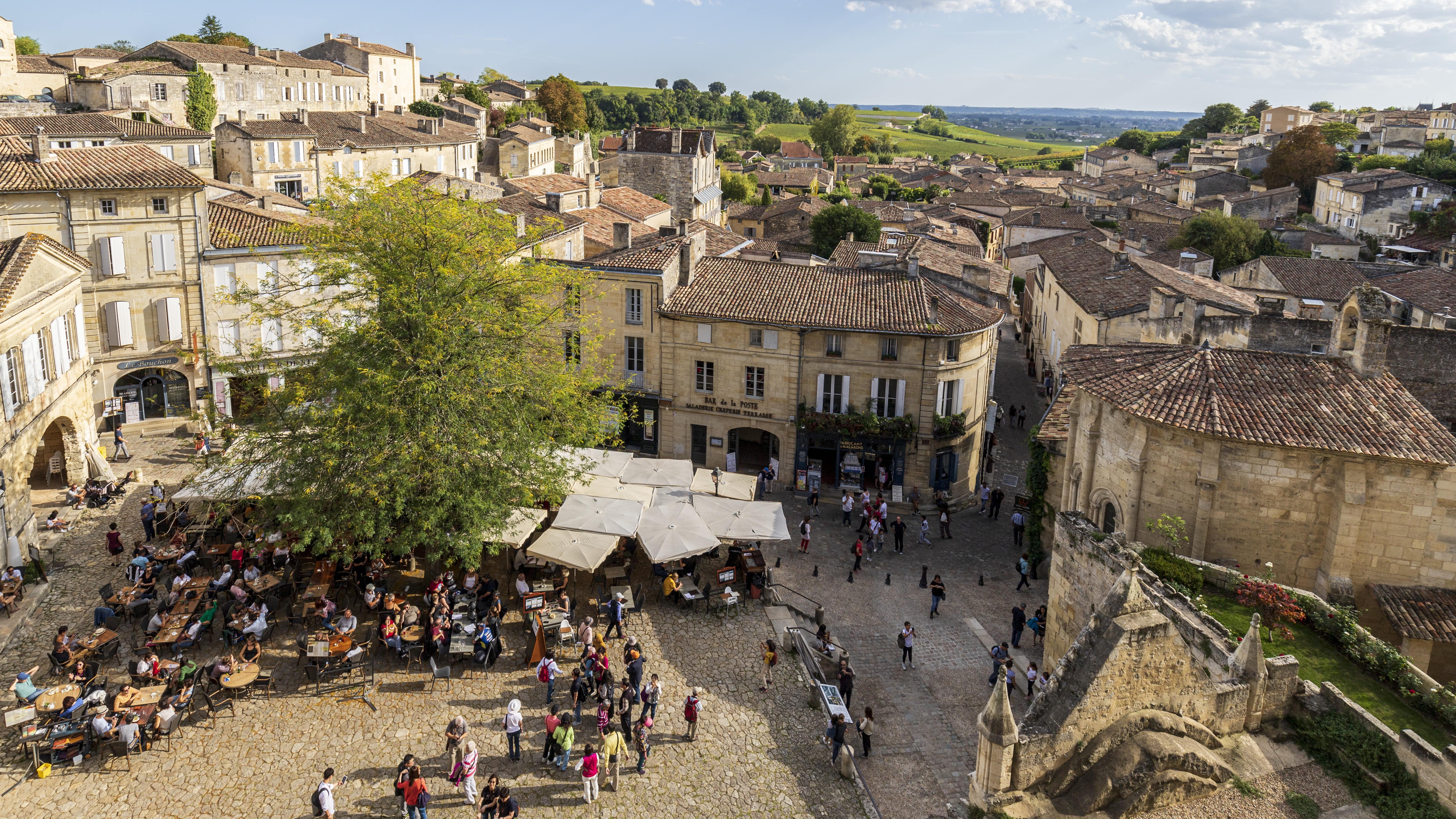 Vue sur la place animée de l’église monolithe de Saint-Émilion depuis son clocher : terrasses de restaurants et badauds