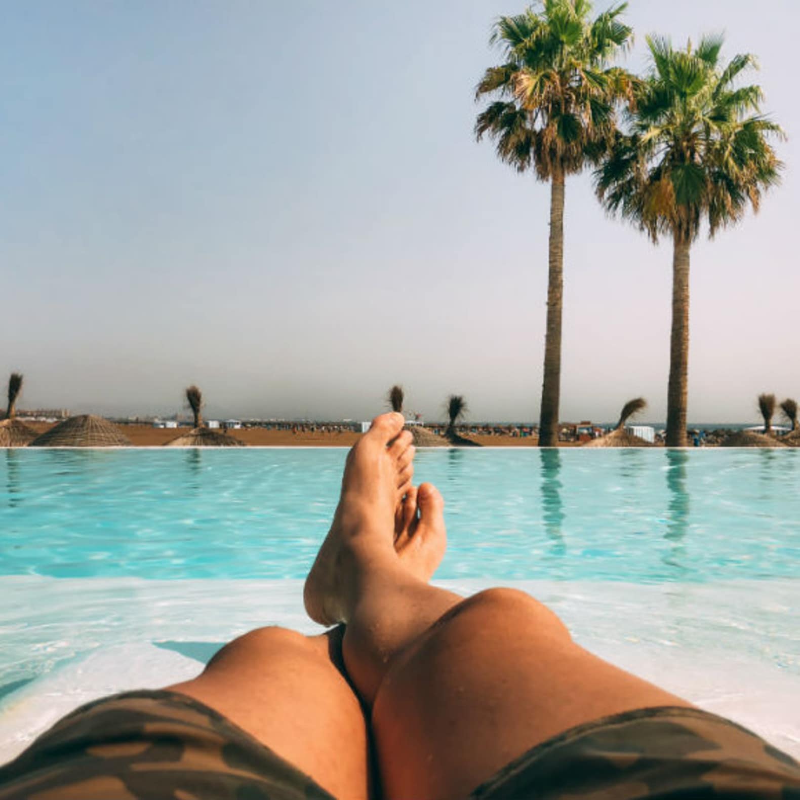 Jambes d’homme allongé devant une piscine à débordement, plage au loin, location maison piscine Espagne
