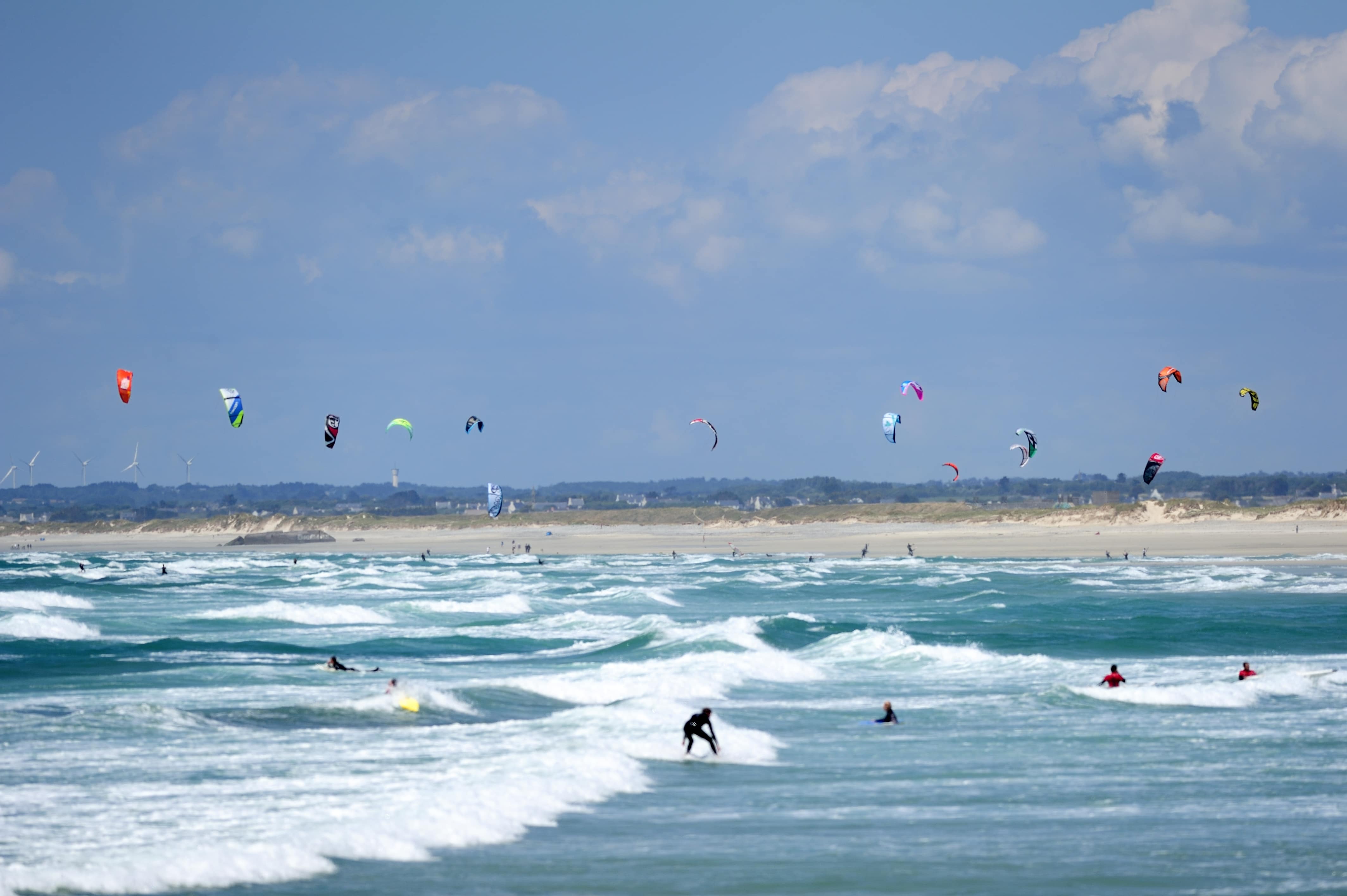 Vue sur la mer, des surfeurs surfent sur les vagues, et sur la plage au loin, au-dessus de laquelle volent des cerfs-volants