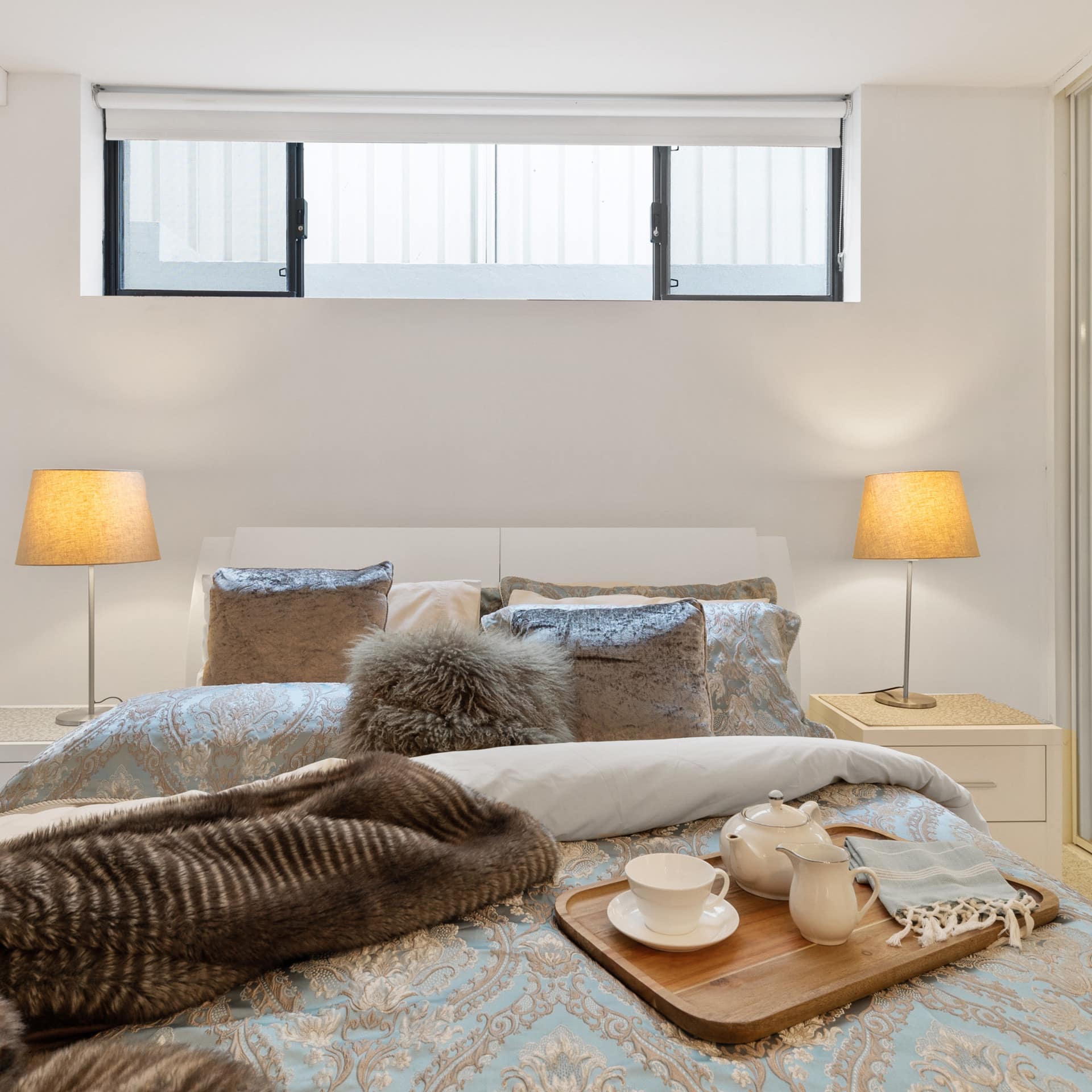 Bedroom with white walls, a queen size bed, with blue floral bedding, faux fur blanket, and tray set with tea on the bed.