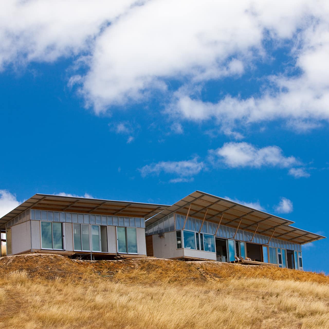 Eco-lodge at Kangaroo Beach Cove with lots of windows on headland with blue sky above and grassy hill in foreground