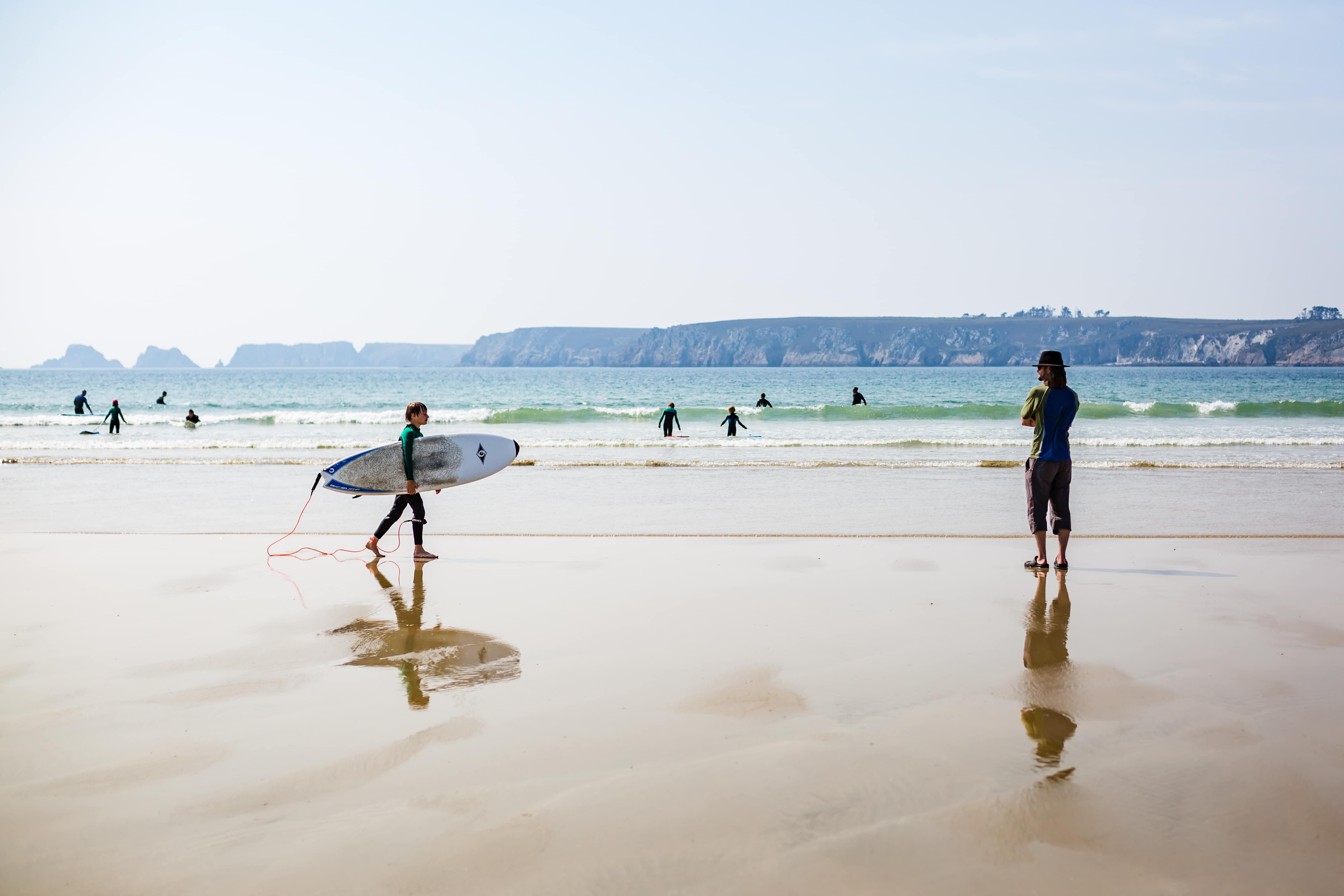 Enfant sur la plage tenant une planche de surf, quelques personnes surfant non loin