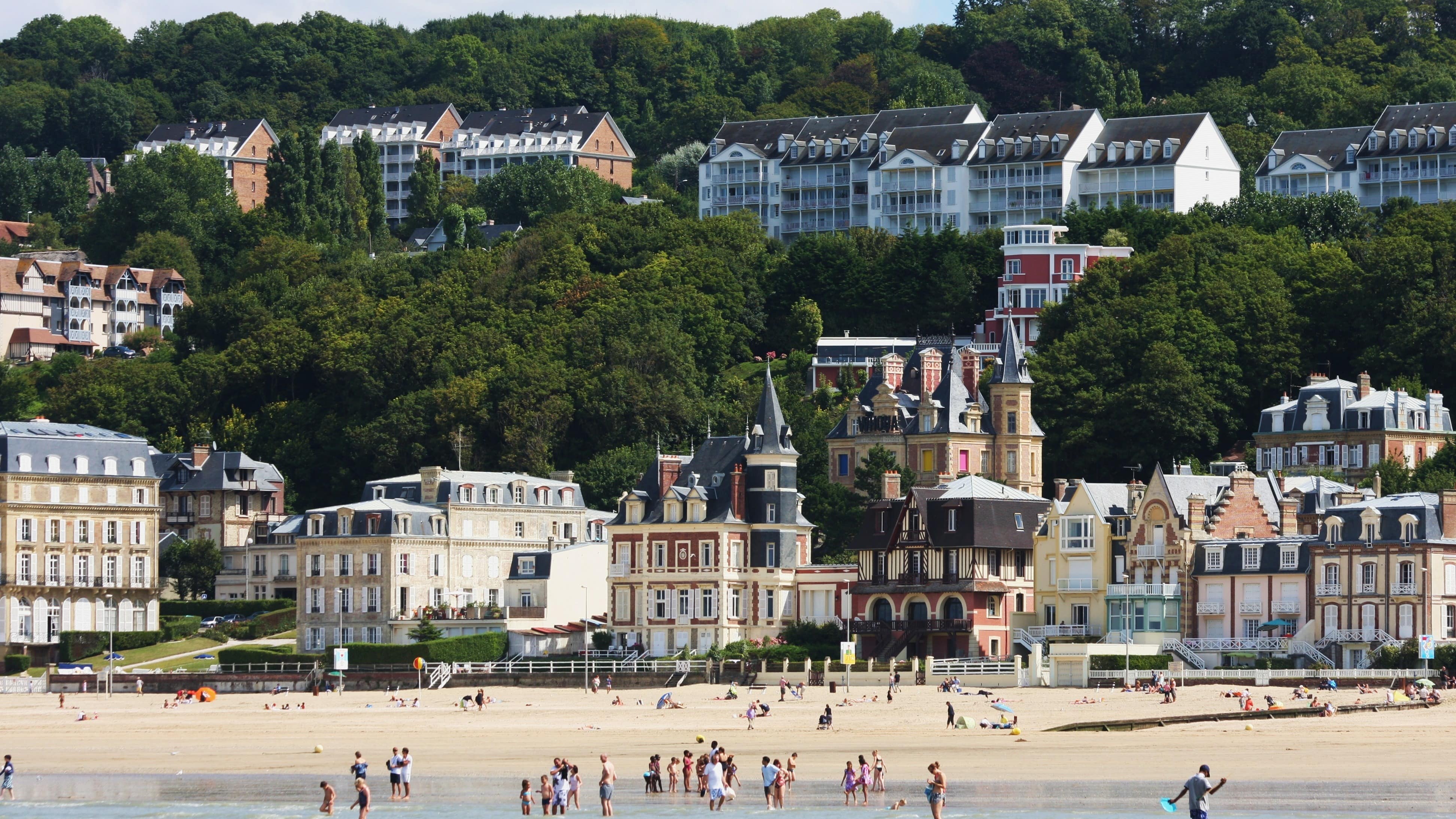 Des personnes se baignent sur la grande plage de sable de Trouville, devant des bâtisses normandes et une colline verdoyante