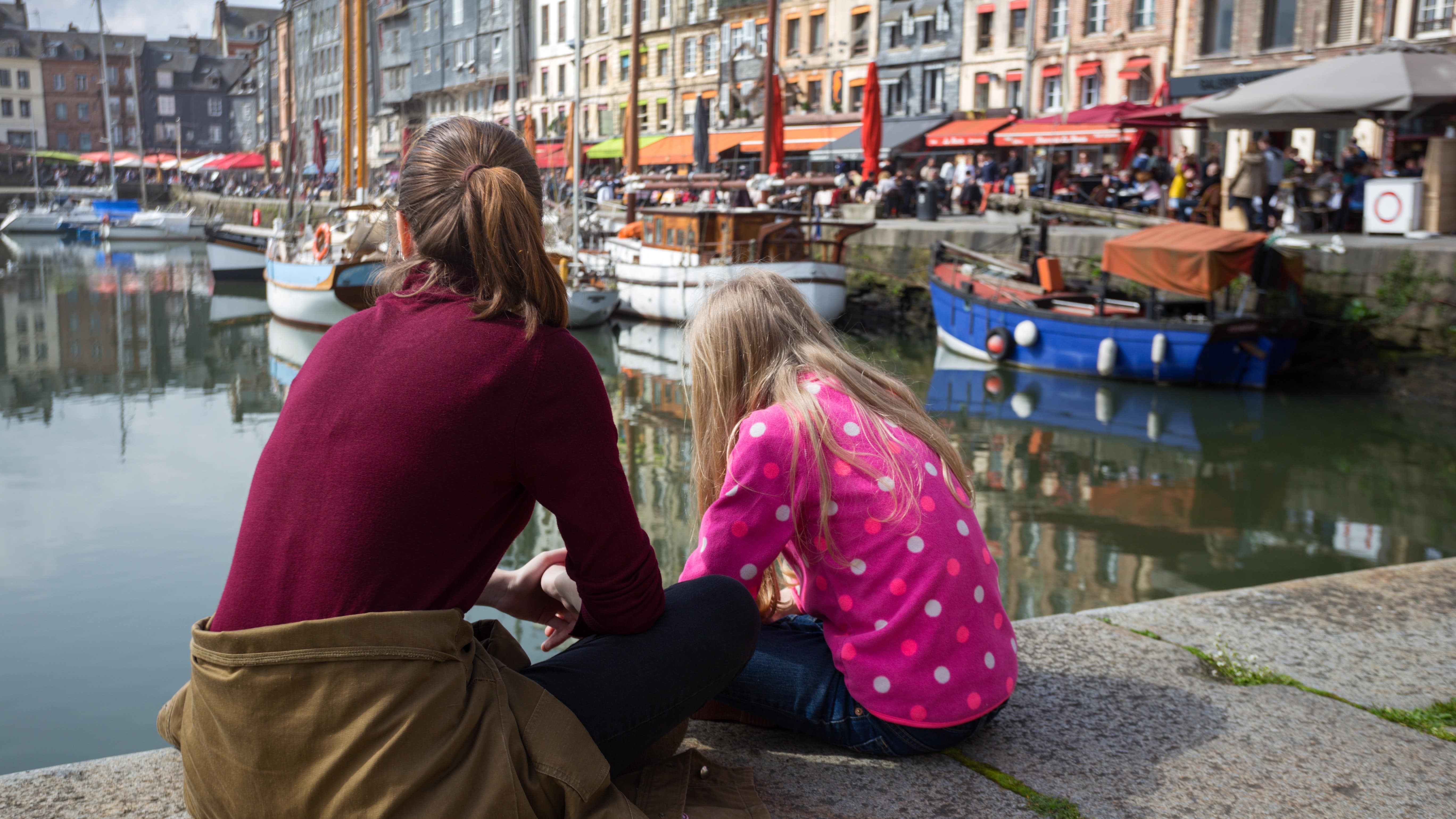 Deux filles assises sur le quai du Vieux Bassin d’Honfleur où sont amarrés des bateaux devant les terrasses des restaurants