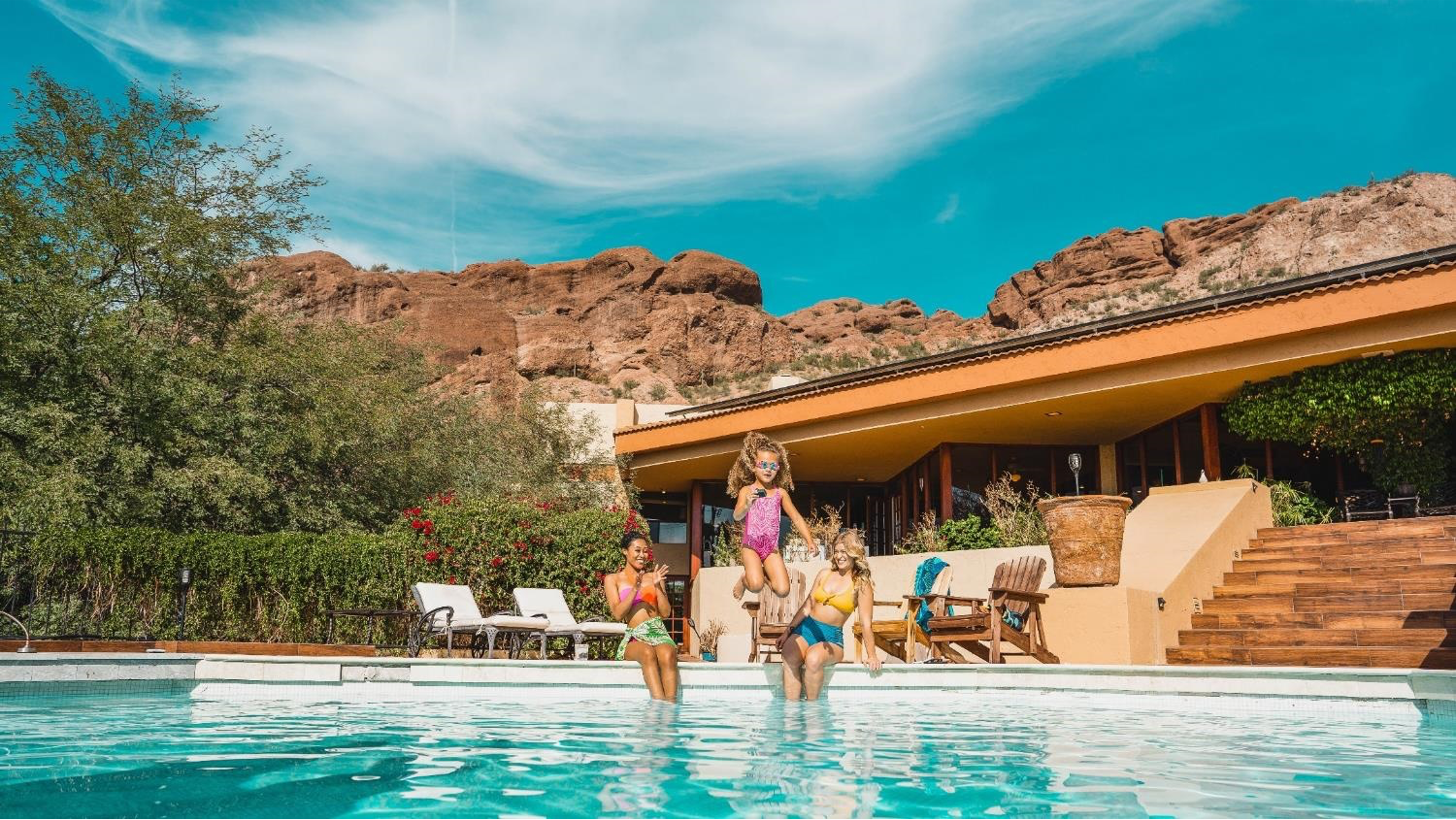 deux femmes assises au bord d'une piscine tandis qu'une petite fille saute dans l'eau entre elles. Il y a des falaises rouges en arrière-plan