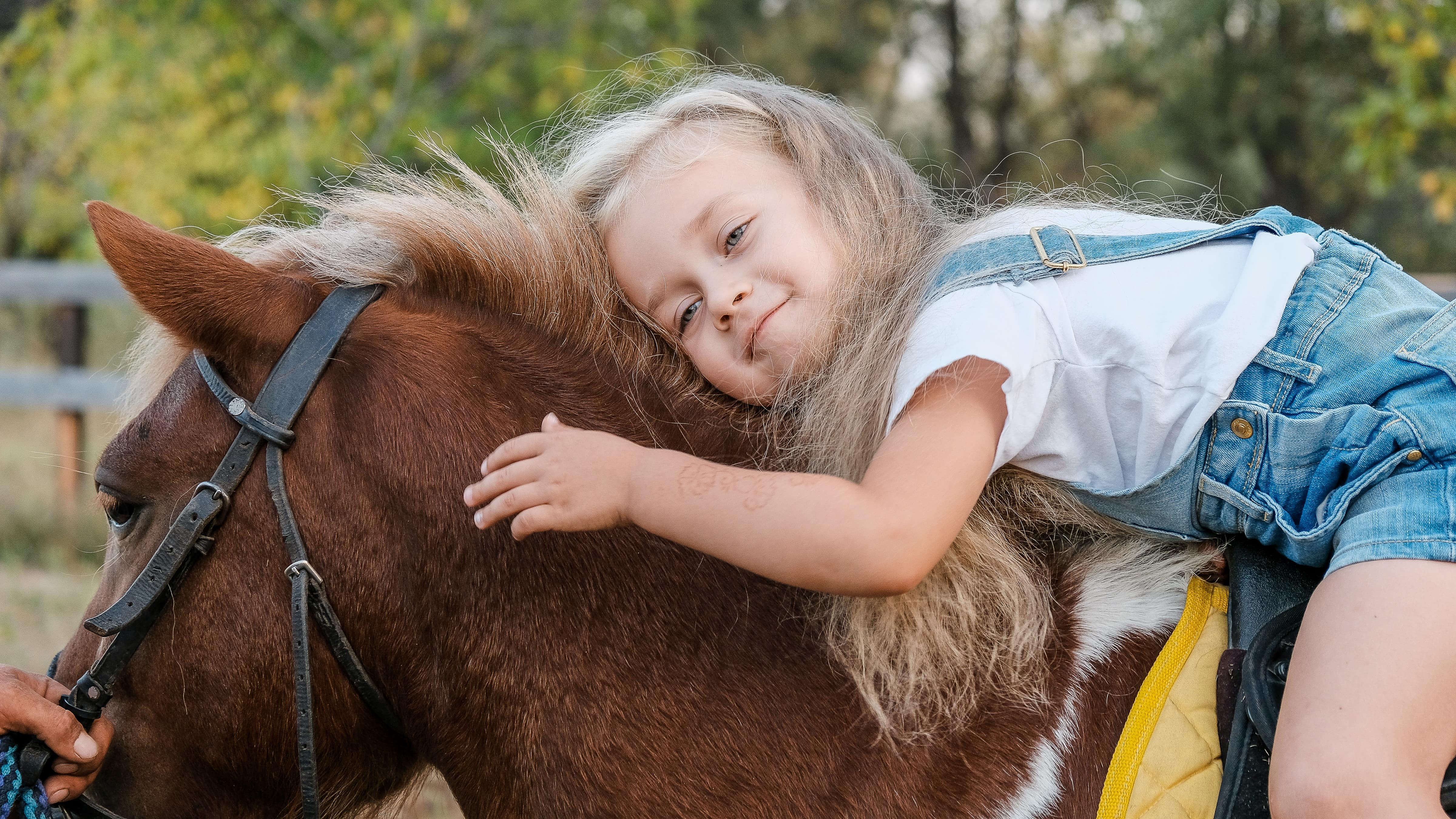 Une petite fille blonde sur un poney, penchée vers sa tête pour lui faire un câlin