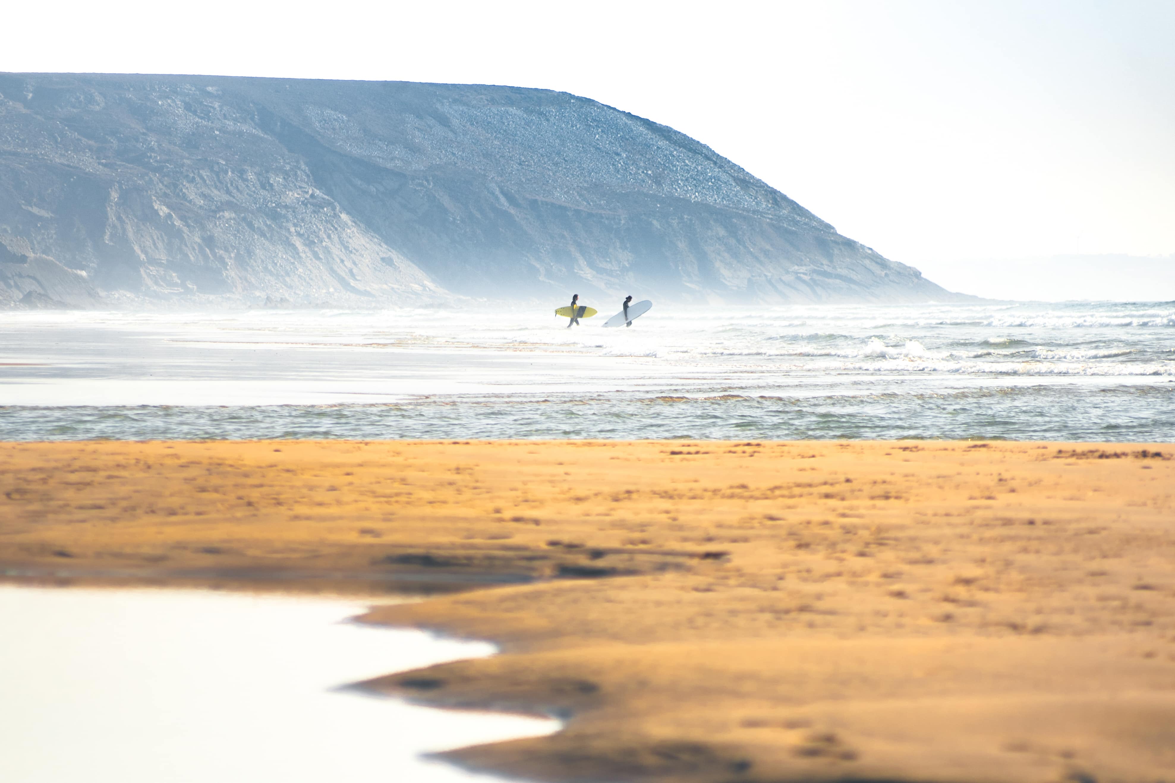 Vue sur la plage, deux surfeurs et des falaises au loin