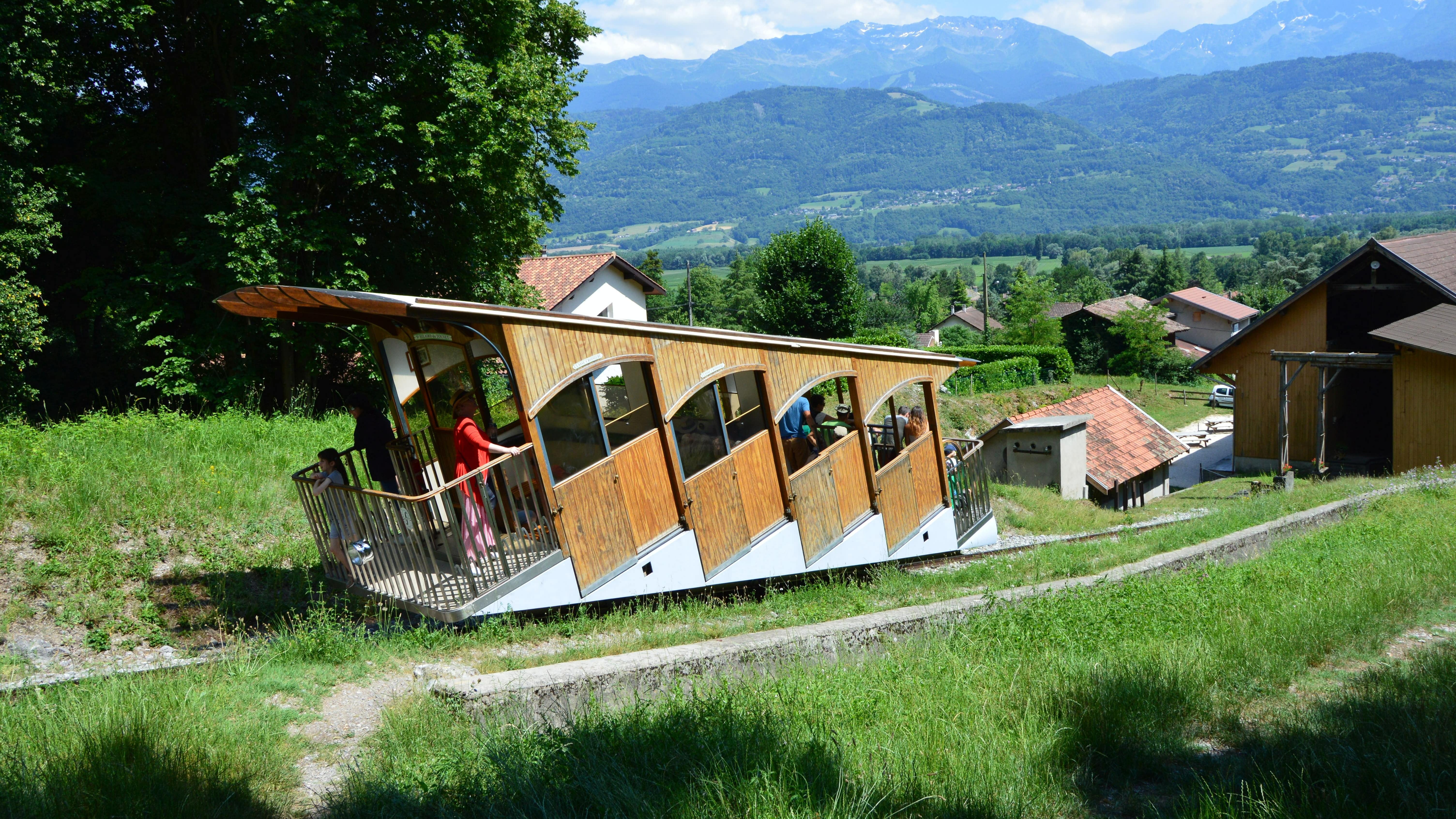 Quelques passagers dans la cabine en bois du funiculaire qui monte la pente herbeuse, avec les montagnes en arrière-plan