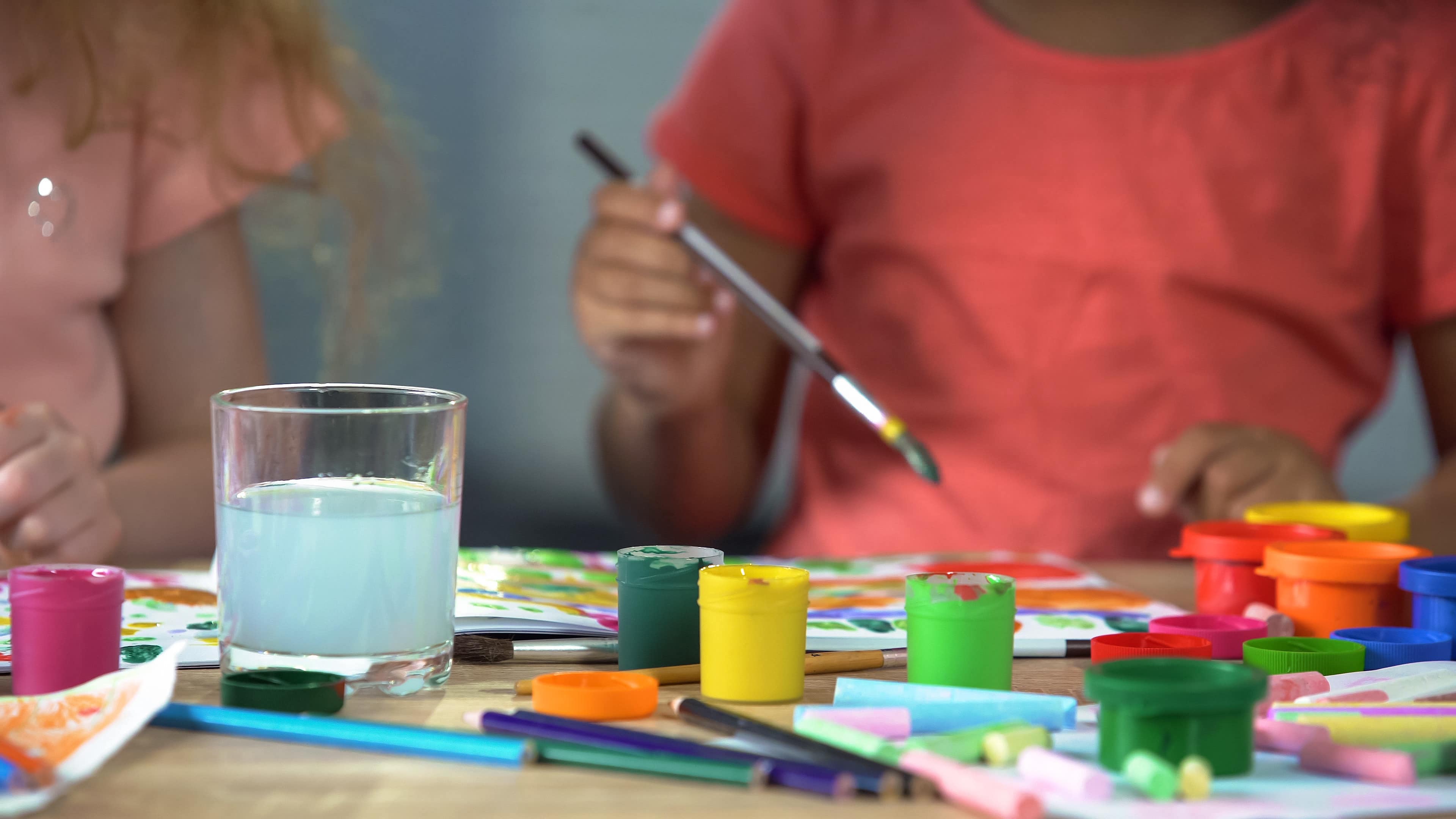 Des enfants en train de peindre sur une table remplie de crayons, craies et pots de peinture