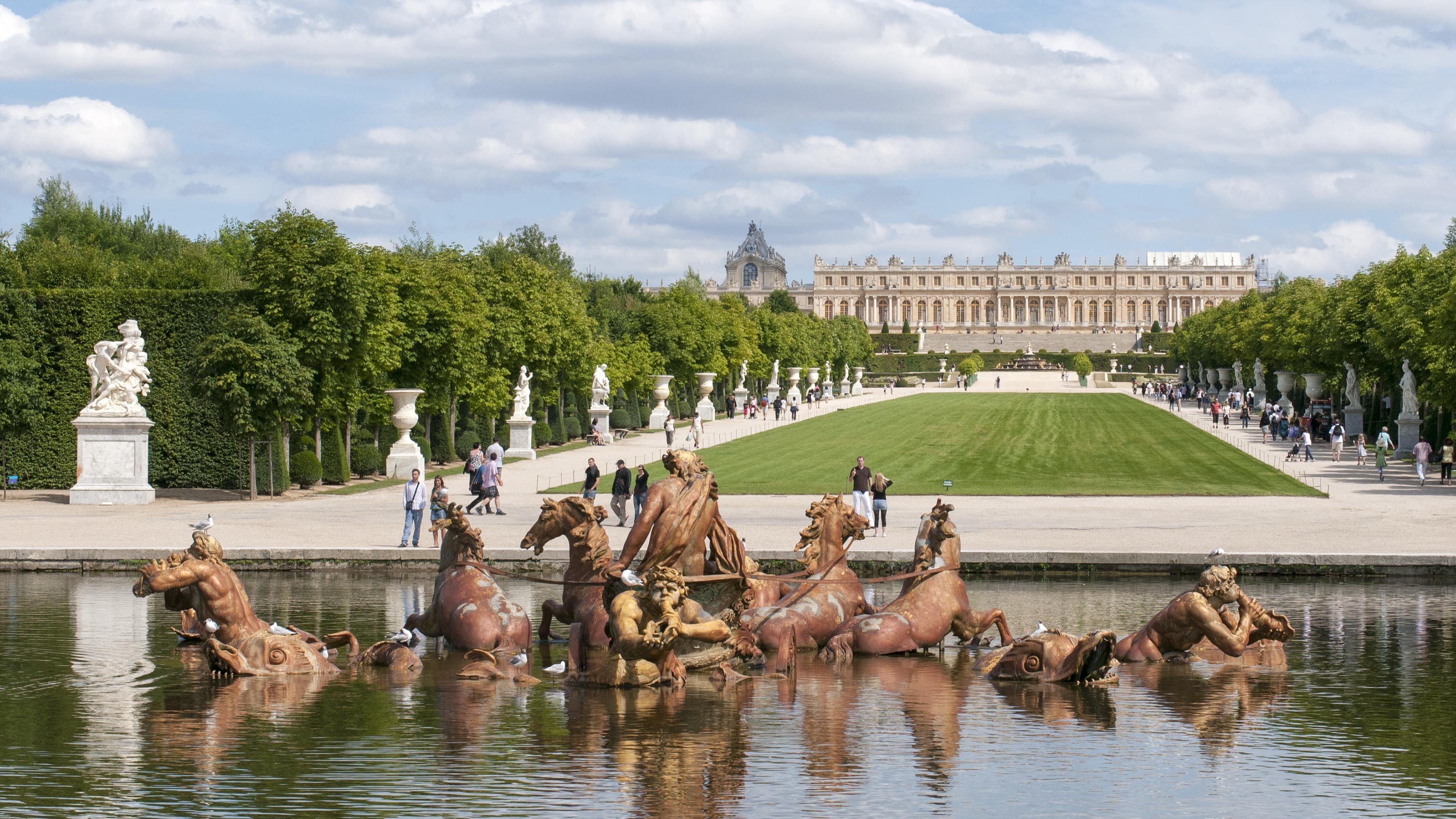 Vue du château de Versailles avec le bassin d’Apollon au premier plan et des gens qui se promènent dans les allées du parc