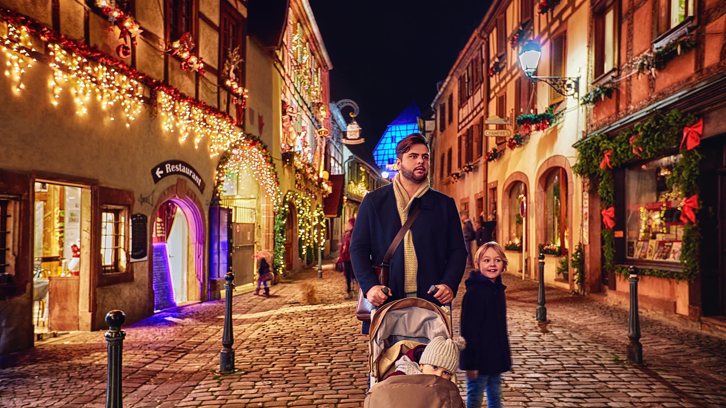 Un père avec deux enfants, dont un en poussette, dans les rues pavées de Kaysersberg décorées pour Noël
