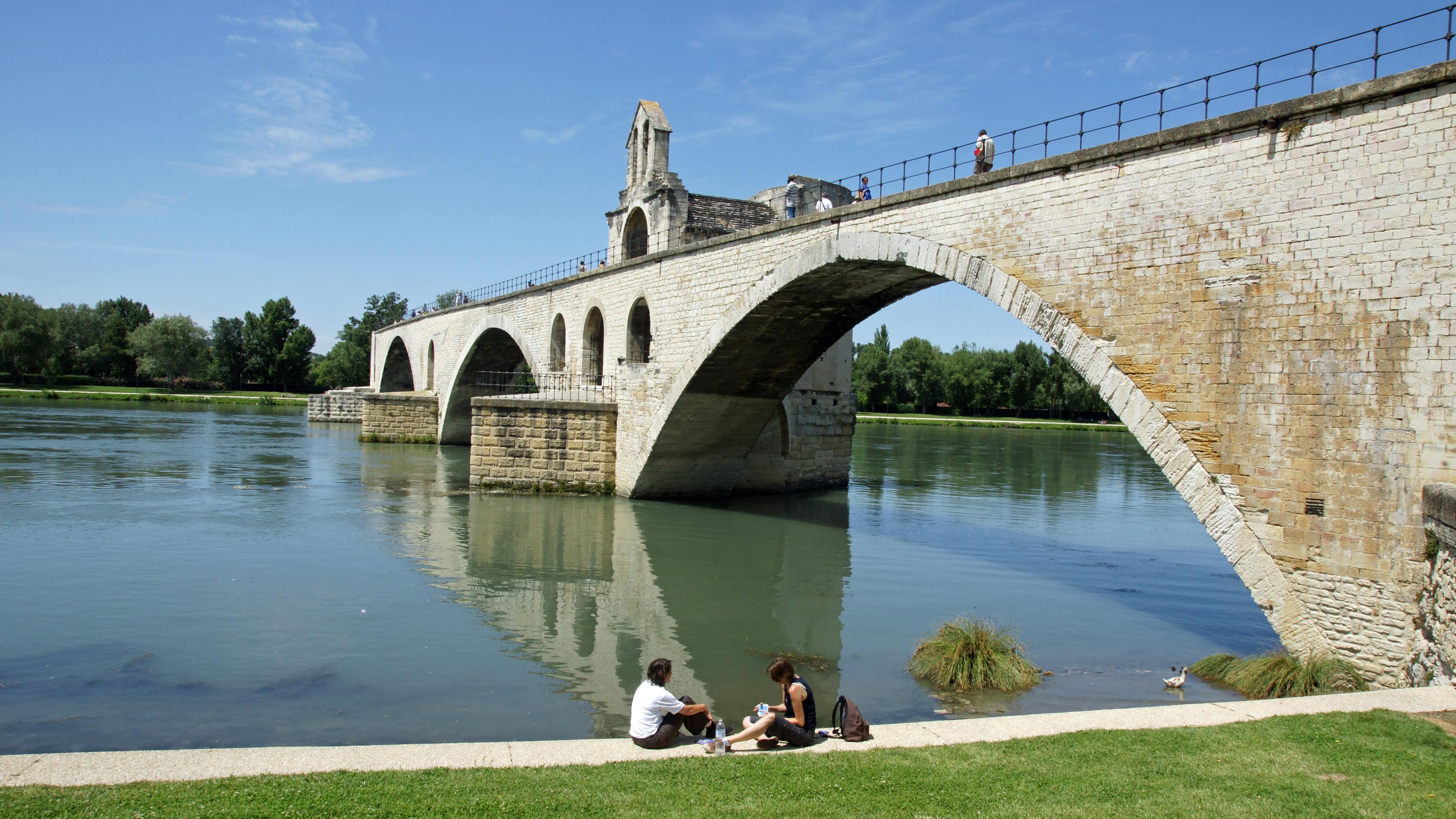 Deux personnes assises sur les rives du Rhône, à côté du pont d’Avignon sur lequel se baladent quelques promeneurs