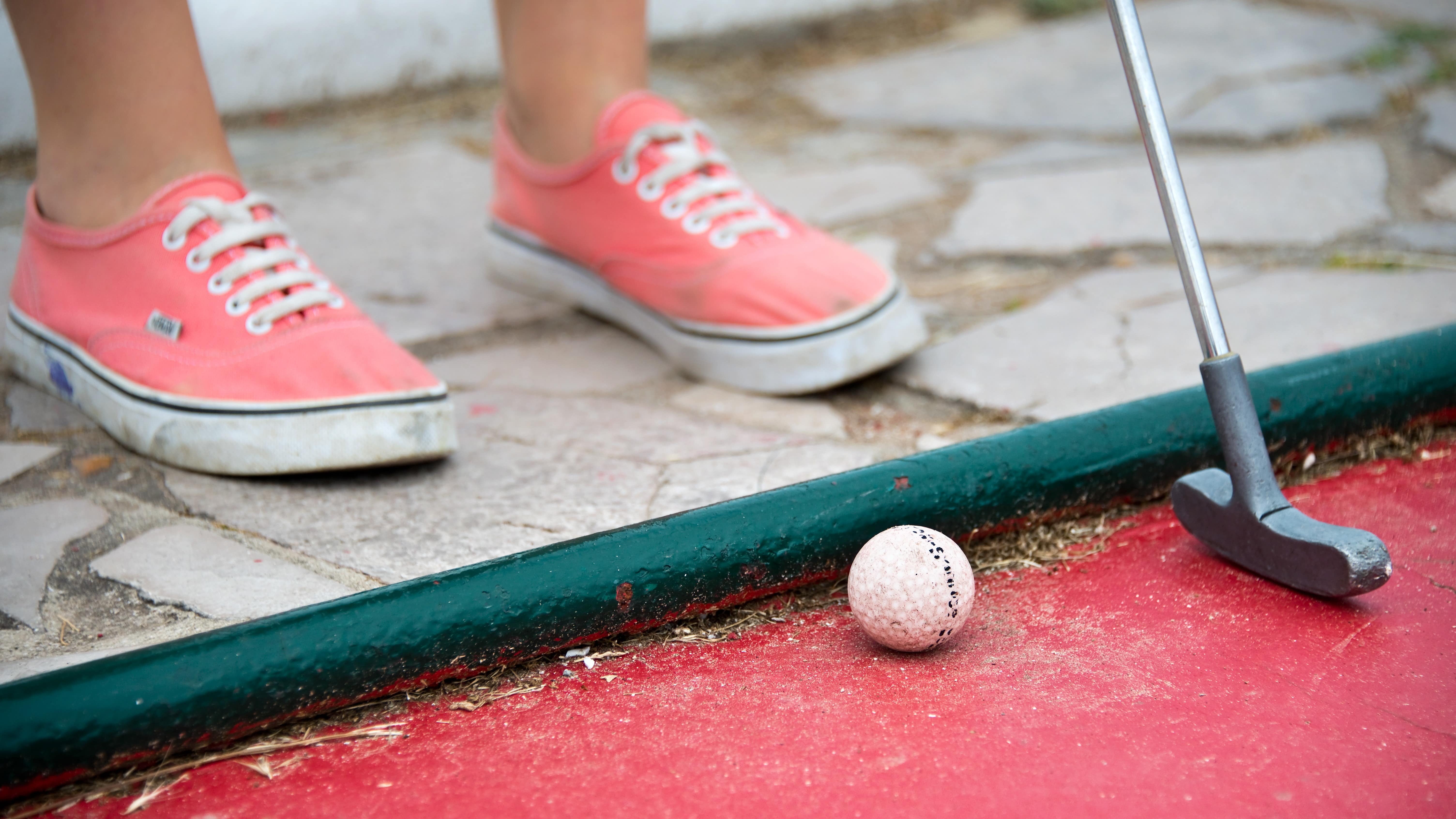 Gros plan sur les chaussures roses d’un enfant qui tient un club de mini-golf, prêt à frapper dans la balle sur le parcours