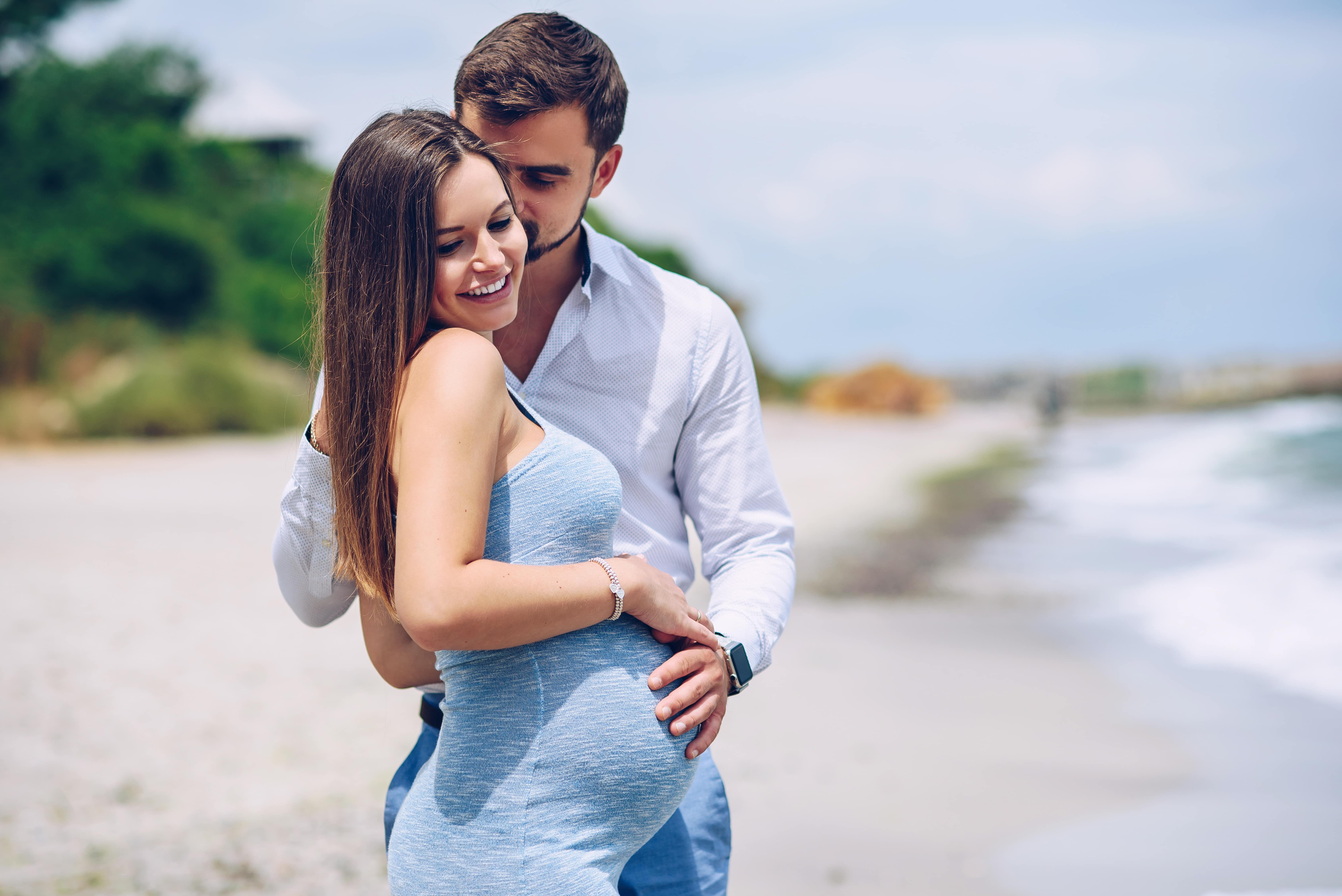 bel homme dans une chemise blanche et un pantalon bleu câlins et embrassant sur une joue sa belle femme enceinte dans une robe bleue sur le fond de la mer.