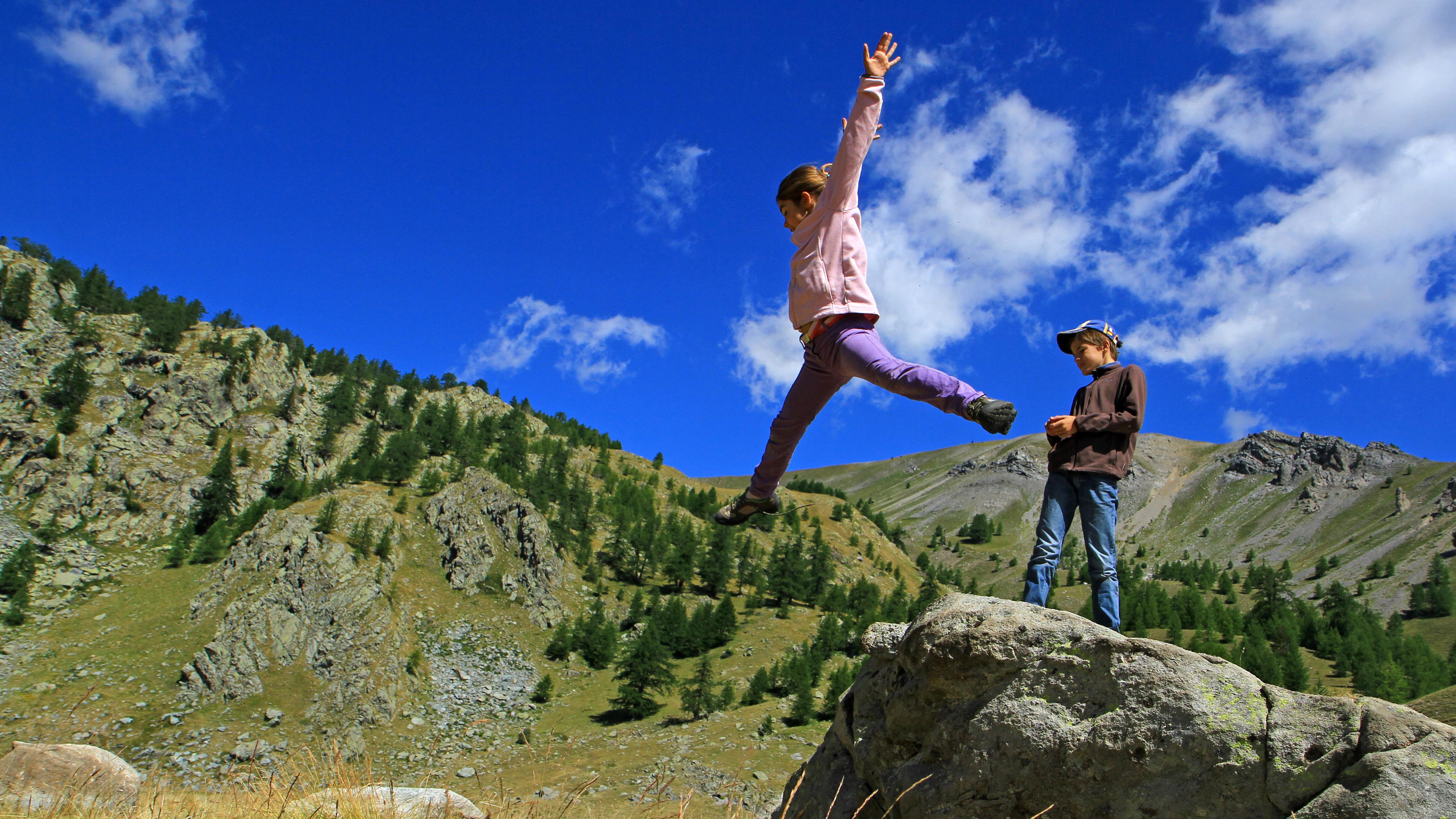 Deux enfants en randonnée dans le Mercantour : la fillette saute depuis un rocher, tandis que le garçon reste sur le rocher.